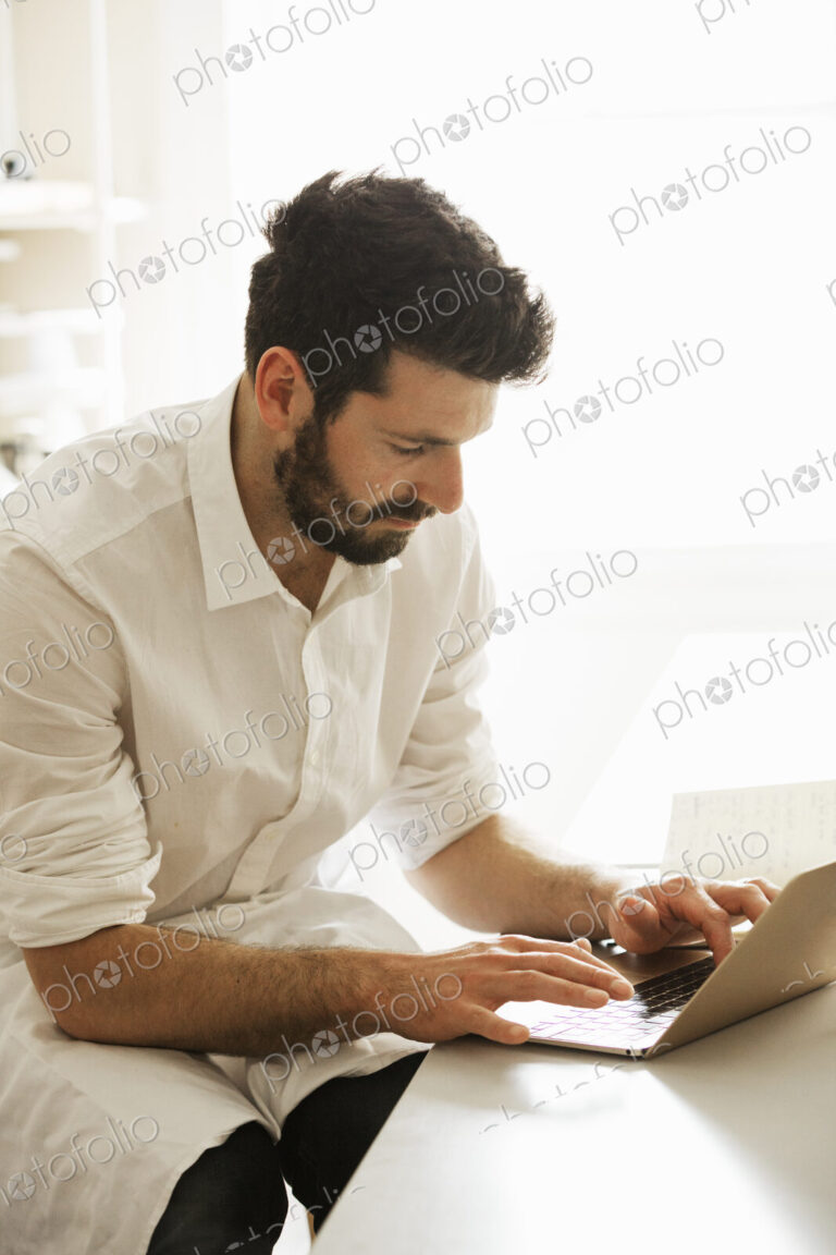 Baker wearing an apron sitting at a laptop computer, typing.