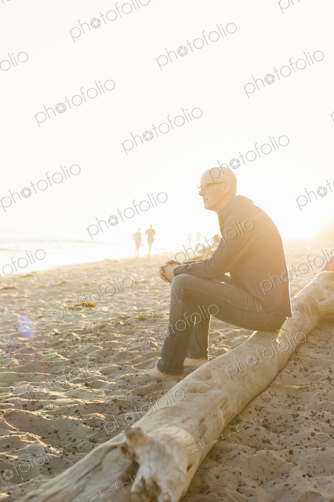 Bald man sitting on a log on a sandy beach.