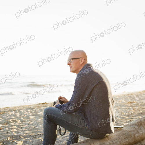 Bald man sitting on a log on a sandy beach. Bald man sitting on a log on a sandy beach.