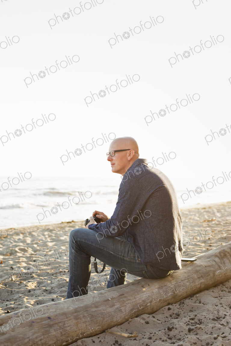 Bald man sitting on a log on a sandy beach.