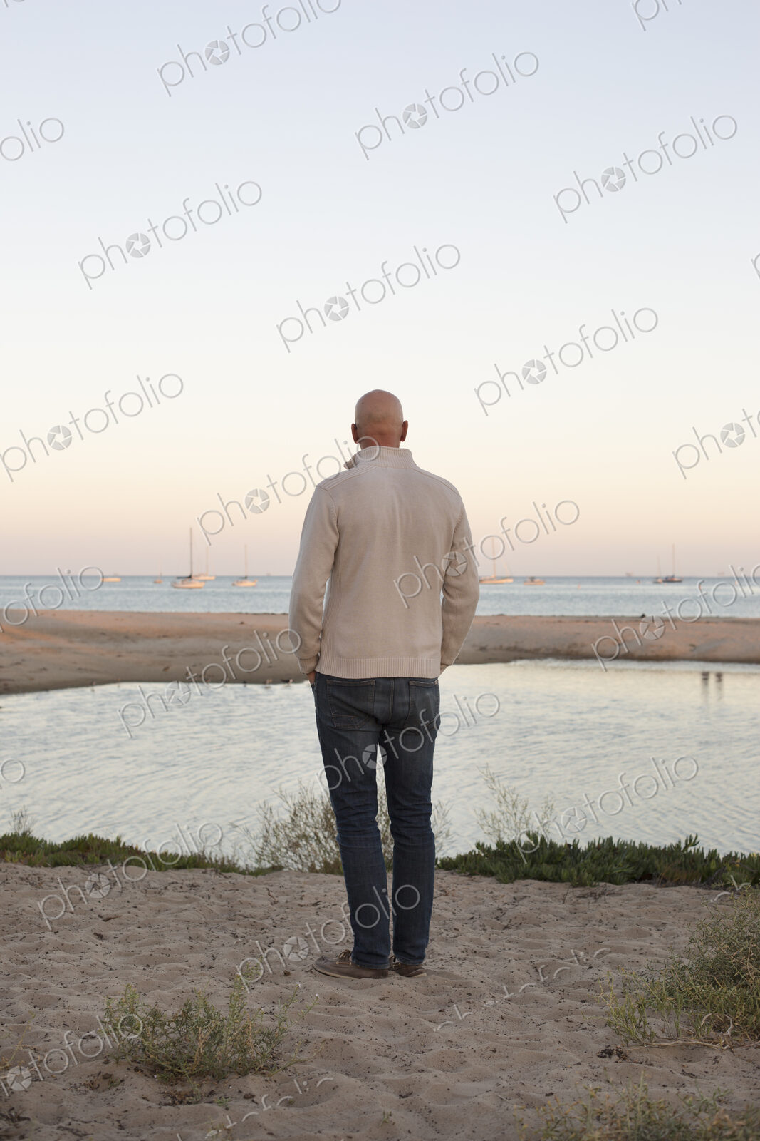 Bald man standing on a sandy beach by the ocean.