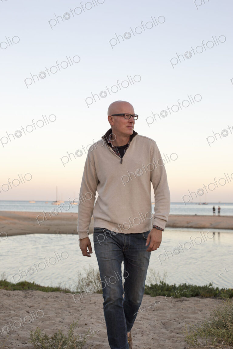 Bald man walking on a sandy beach by the ocean.