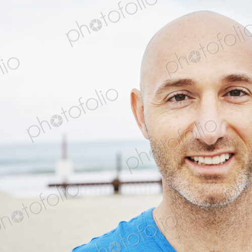 Bald mature man standing on a beach, smiling at camera.