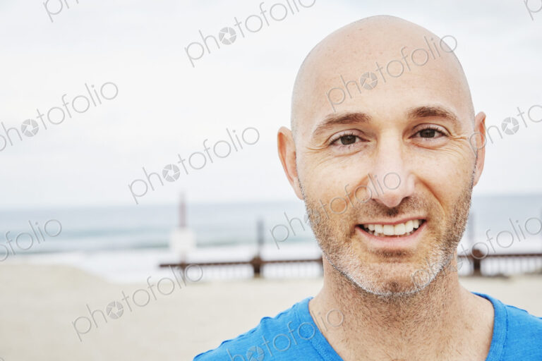 Bald mature man standing on a beach, smiling at camera.