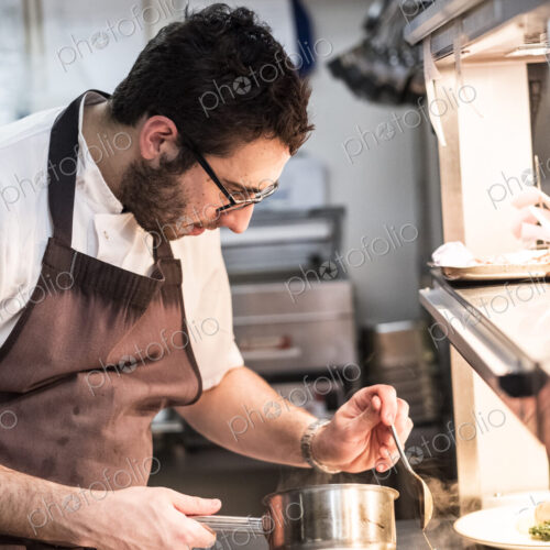 Bearded chef wearing glasses and apron standing in commercial kitchen at counter,…