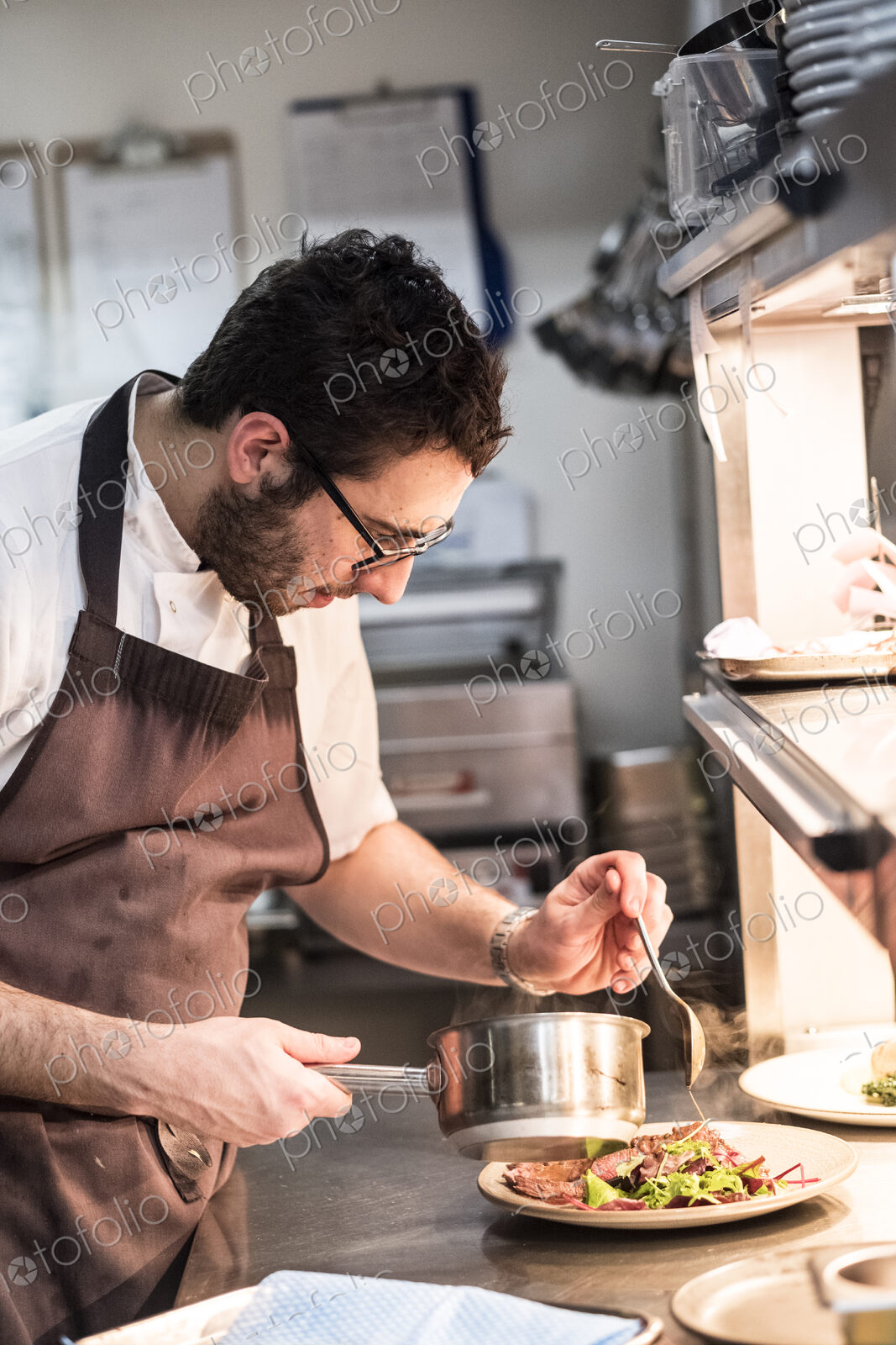 Bearded chef wearing glasses and apron standing in commercial kitchen at counter,…