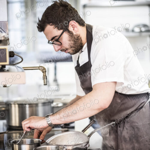 Bearded chef wearing glasses and apron standing in commercial kitchen at stove,…