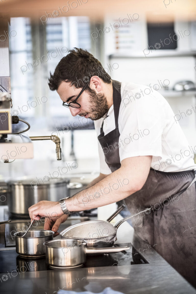 Bearded chef wearing glasses and apron standing in commercial kitchen at stove,…