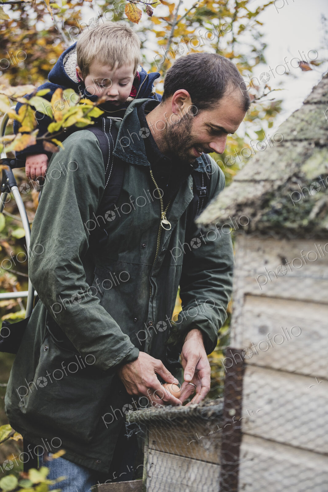 Bearded man carrying young boy on his back picking fresh eggs from chicken coop on a farm.
