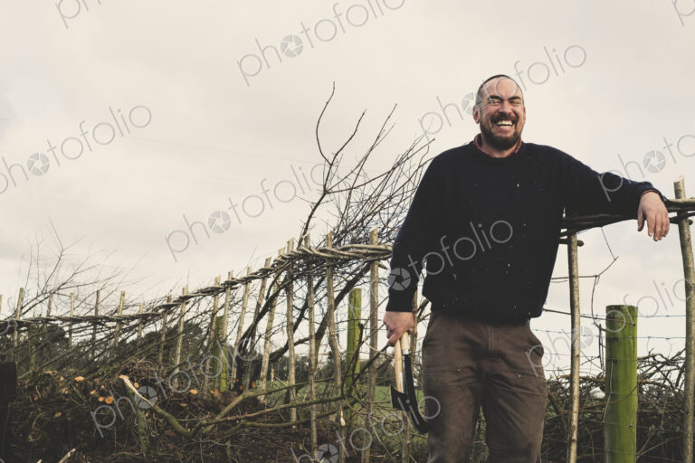 Bearded man holding axe and bill hook standing next to a newly built traditional…