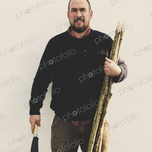 Bearded man holding bill hook and bunch of wooden stakes for traditional hedge laying,…