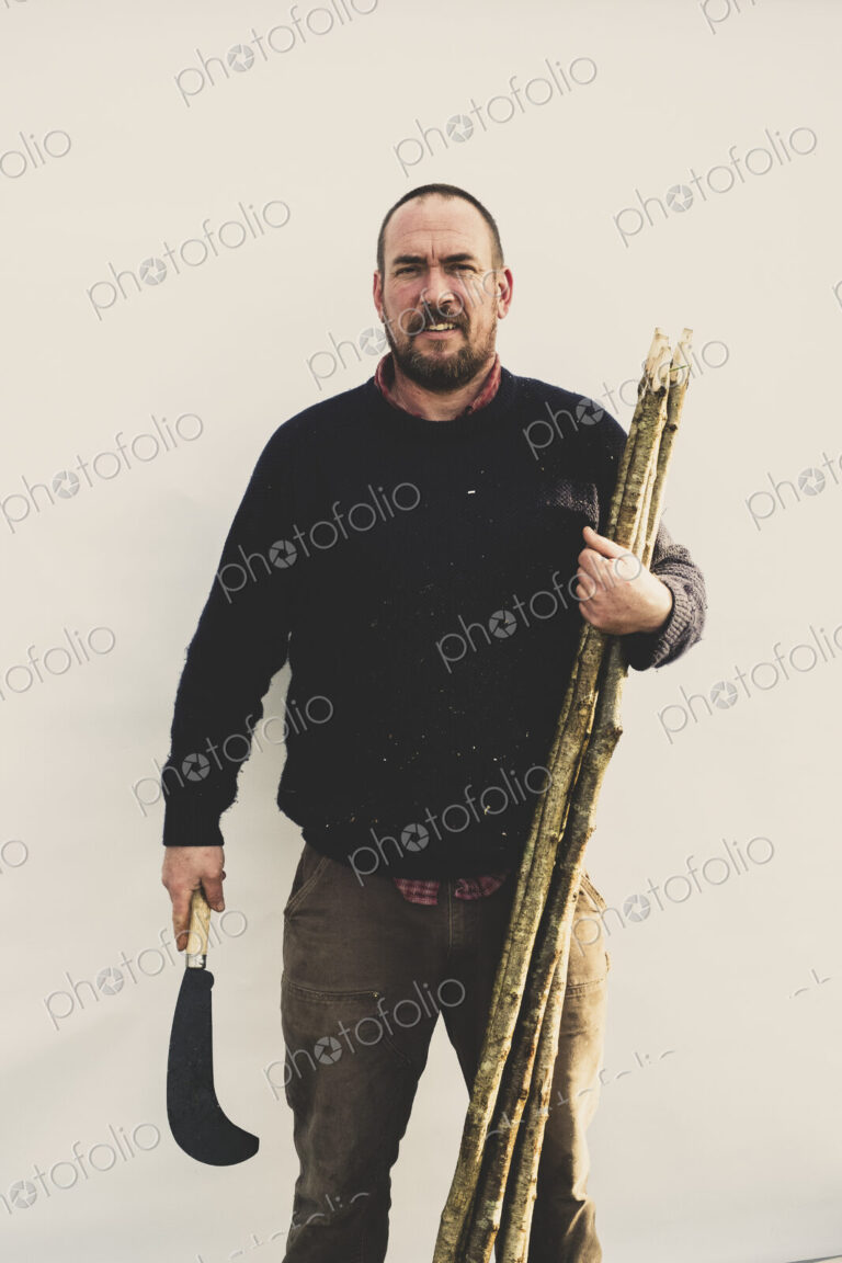 Bearded man holding bill hook and bunch of wooden stakes for traditional hedge laying,…