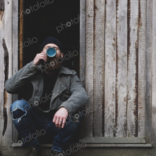 Bearded man sitting in doorway of wooden workshop, drinking tea from blue mug.