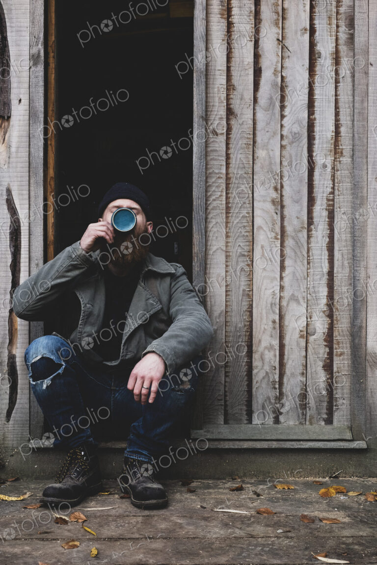 Bearded man sitting in doorway of wooden workshop, drinking tea from blue mug.