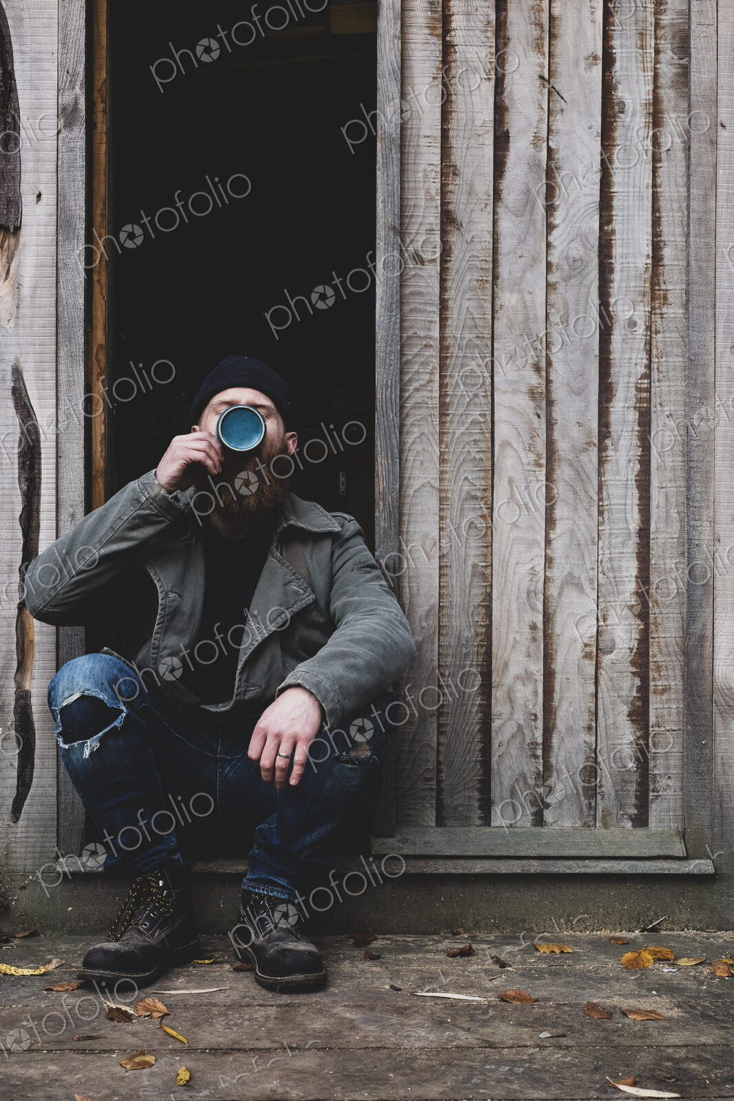 Bearded man sitting in doorway of wooden workshop, drinking tea from blue mug.
