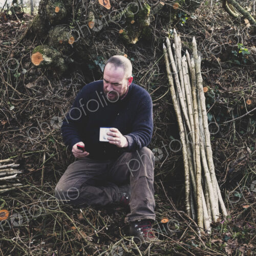 Bearded man sitting on ground next to bunch of wooden stakes, holding mug, checking…