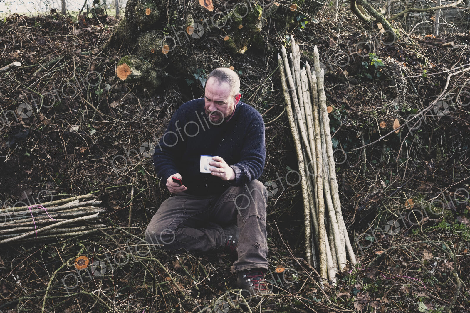 Bearded man sitting on ground next to bunch of wooden stakes, holding mug, checking…
