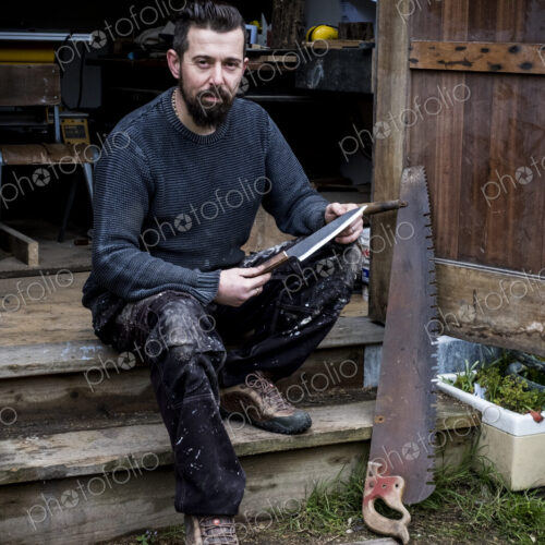 Bearded man sitting on steps outside his workshop, holding handmade knife, looking at… Bearded man sitting on steps outside his workshop, holding handmade knife, looking at…