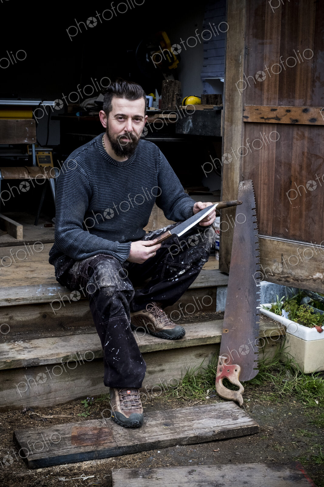 Bearded man sitting on steps outside his workshop, holding handmade knife, looking at…