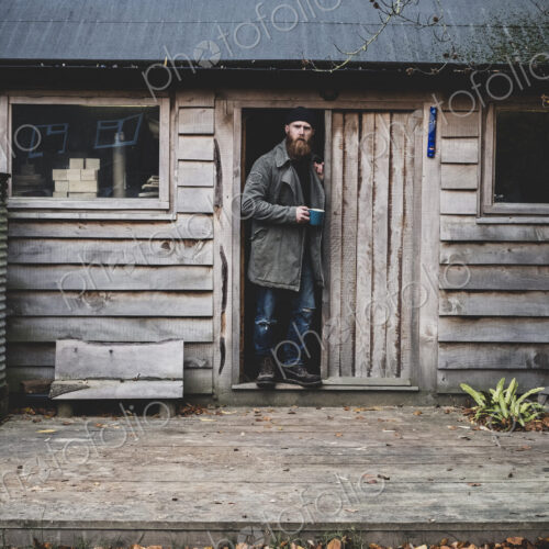 Bearded man standing in doorway of wooden workshop, holding blue mug, looking at camera.