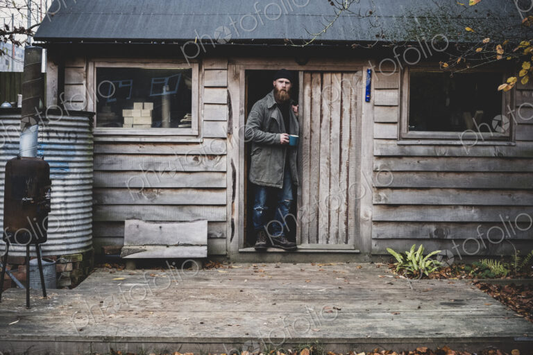 Bearded man standing in doorway of wooden workshop, holding blue mug, looking at camera.