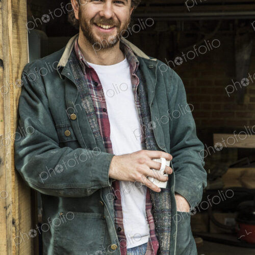 Bearded man standing in doorway of woodworking workshop, holding mug, smiling at camera.