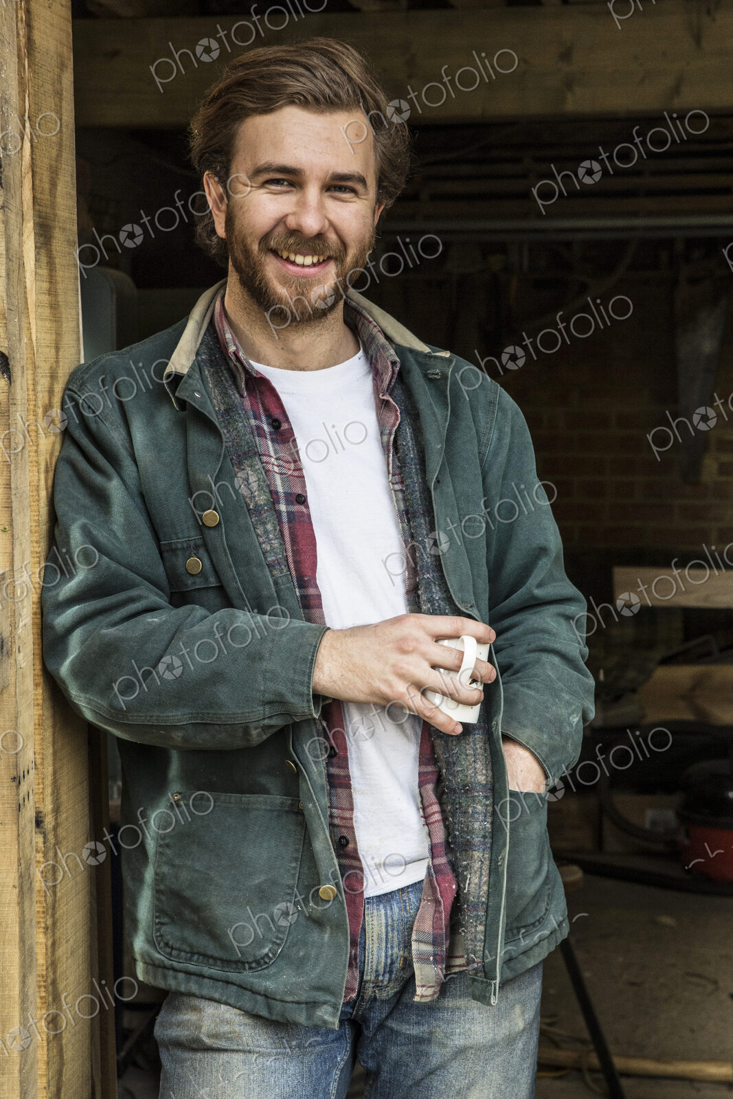 Bearded man standing in doorway of woodworking workshop, holding mug, smiling at camera.