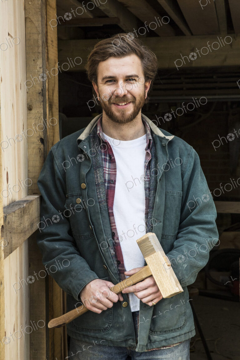 Bearded man standing in doorway of woodworking workshop, holding wooden mallet,…