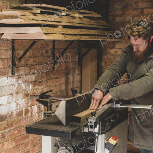 Bearded man standing in workshop, wearing ear protectors, working on piece of wood.