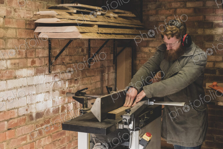 Bearded man standing in workshop, wearing ear protectors, working on piece of wood.
