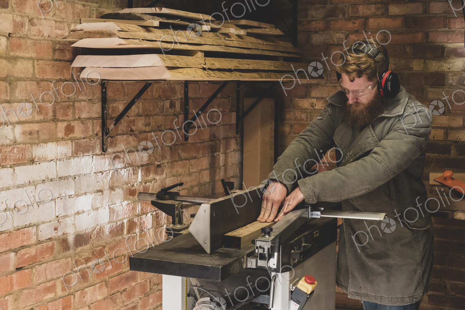 Bearded man standing in workshop, wearing ear protectors, working on piece of wood.