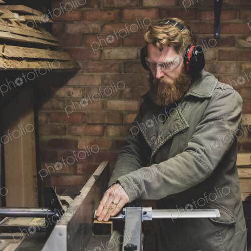 Bearded man standing in workshop, wearing ear protectors, working on piece of wood.