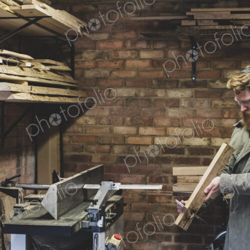 Bearded man standing in workshop, wearing ear protectors, working on piece of wood.