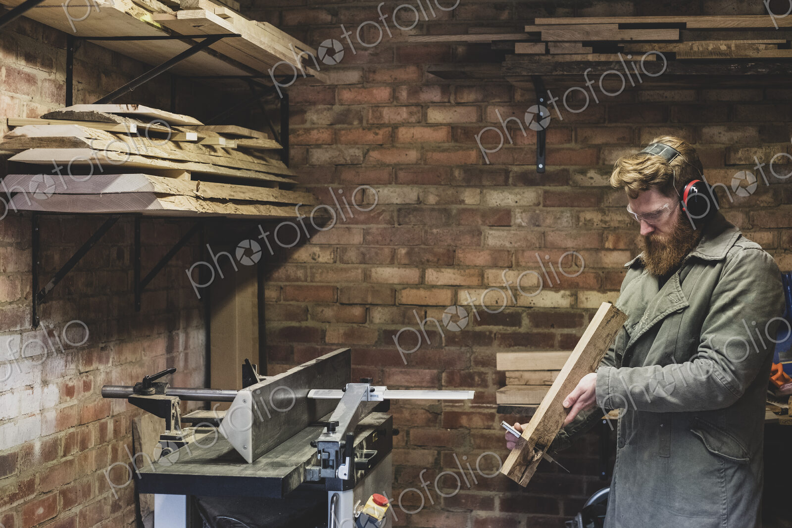 Bearded man standing in workshop, wearing ear protectors, working on piece of wood.