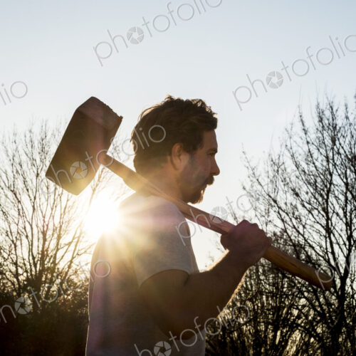 Bearded man walking outdoors at sunset, carrying large wooden mallet on his shoulder. Bearded man walking outdoors at sunset, carrying large wooden mallet on his shoulder.