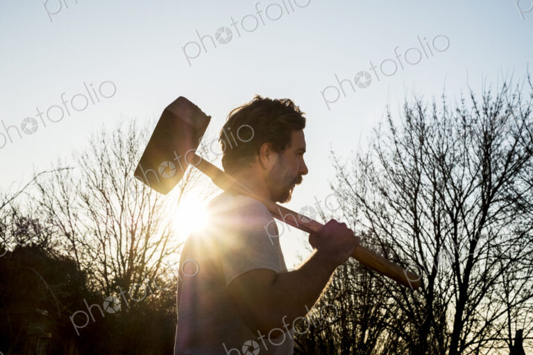 Bearded man walking outdoors at sunset, carrying large wooden mallet on his shoulder.