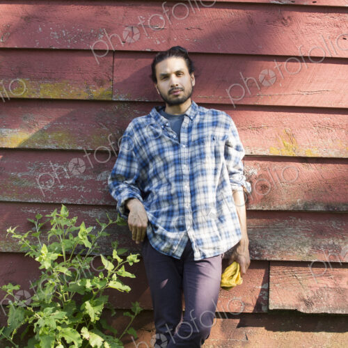Bearded man wearing a checkered shirt leaning against a wooden wall in the shade.