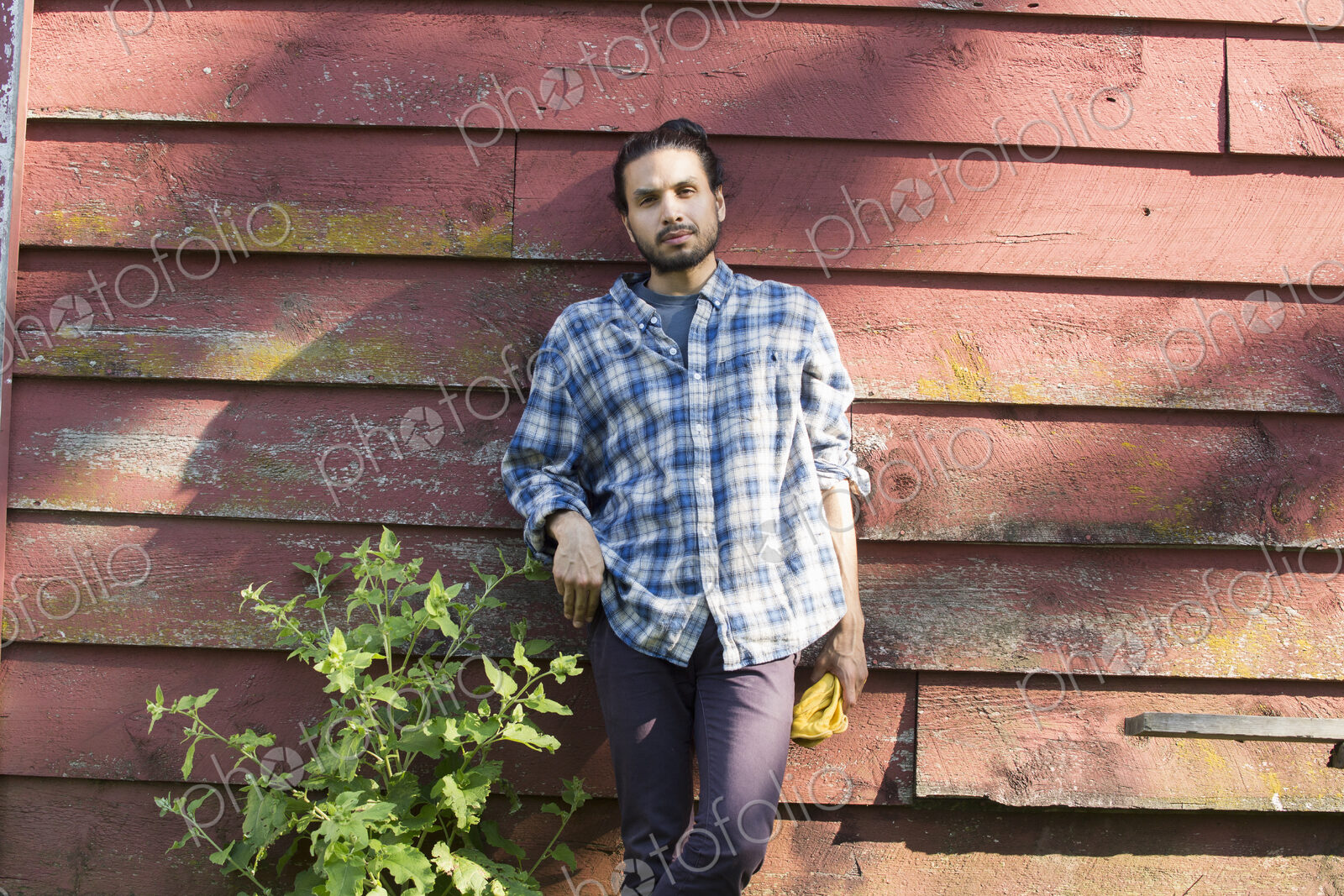 Bearded man wearing a checkered shirt leaning against a wooden wall in the shade.