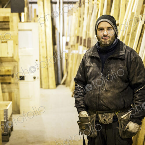 Bearded man wearing beanie and tool belt standing next to a stack of wooden planks in… Bearded man wearing beanie and tool belt standing next to a stack of wooden planks in…
