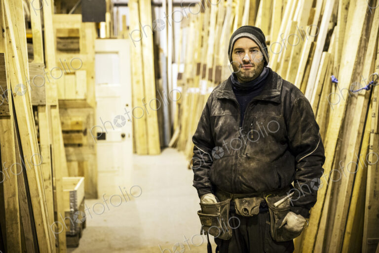 Bearded man wearing beanie and tool belt standing next to a stack of wooden planks in…