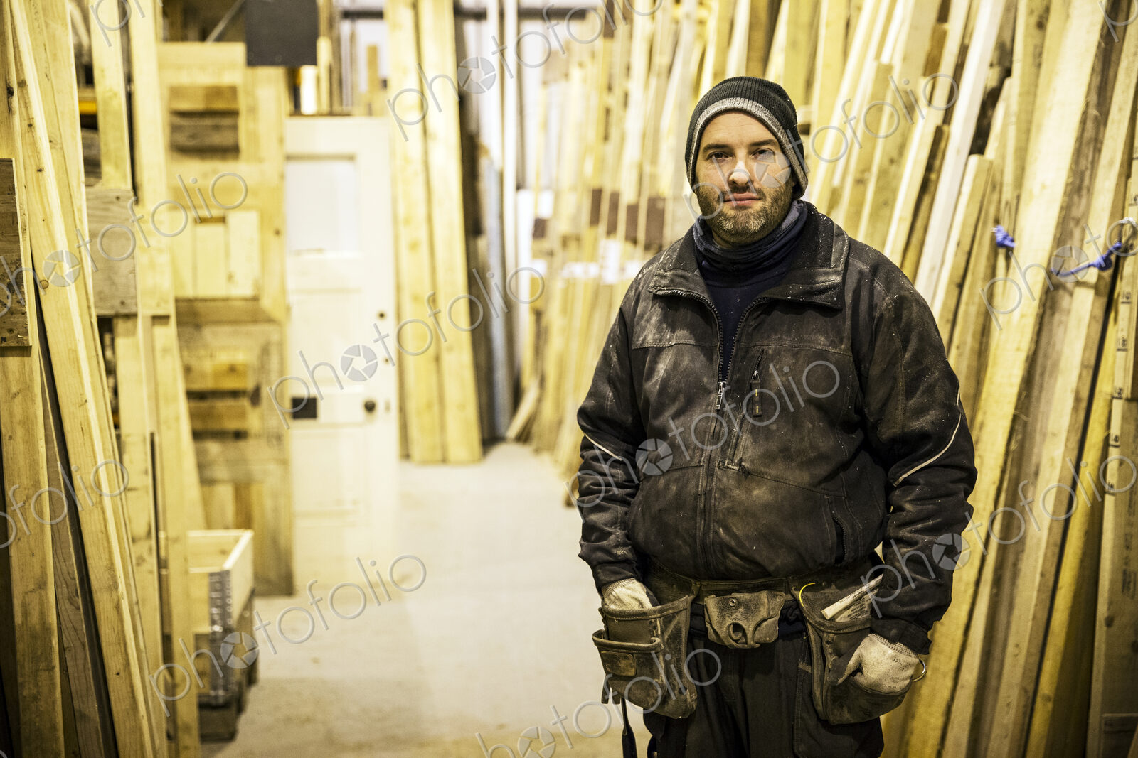 Bearded man wearing beanie and tool belt standing next to a stack of wooden planks in…