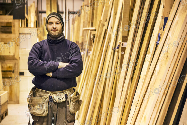 Bearded man wearing beanie and tool belt standing with his arms crossed next to a…