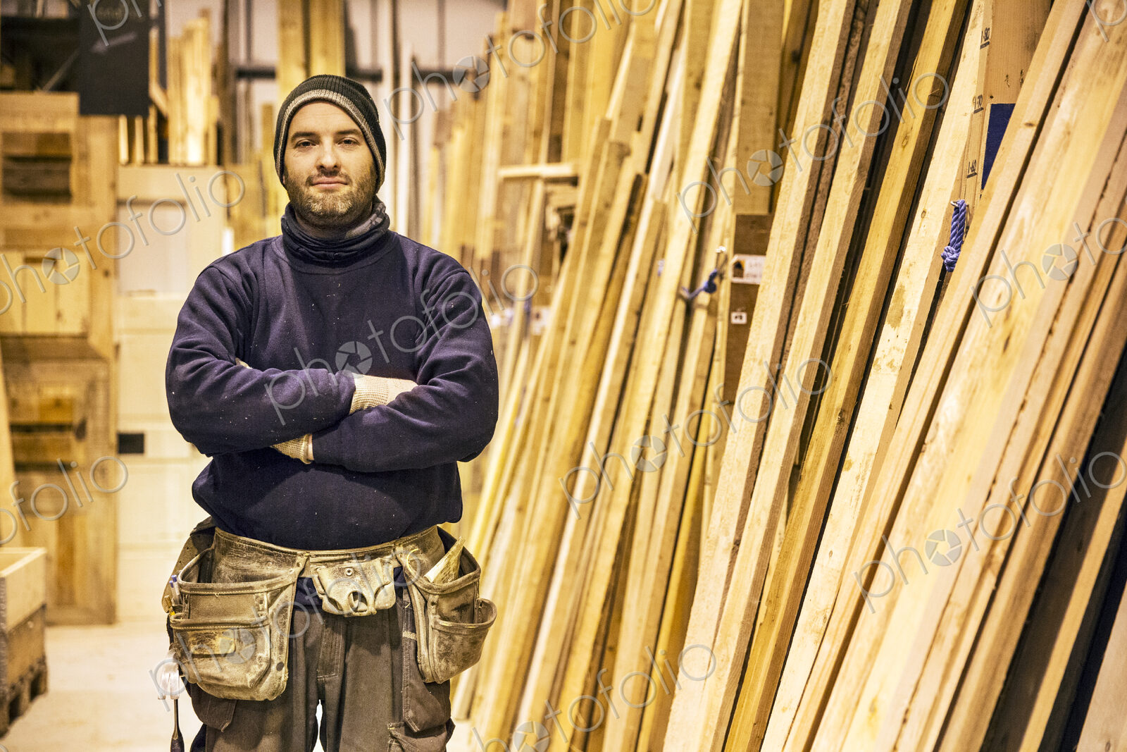 Bearded man wearing beanie and tool belt standing with his arms crossed next to a…