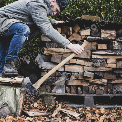 Bearded man wearing black beanie and parka standing in garden in autumn, using axe to…