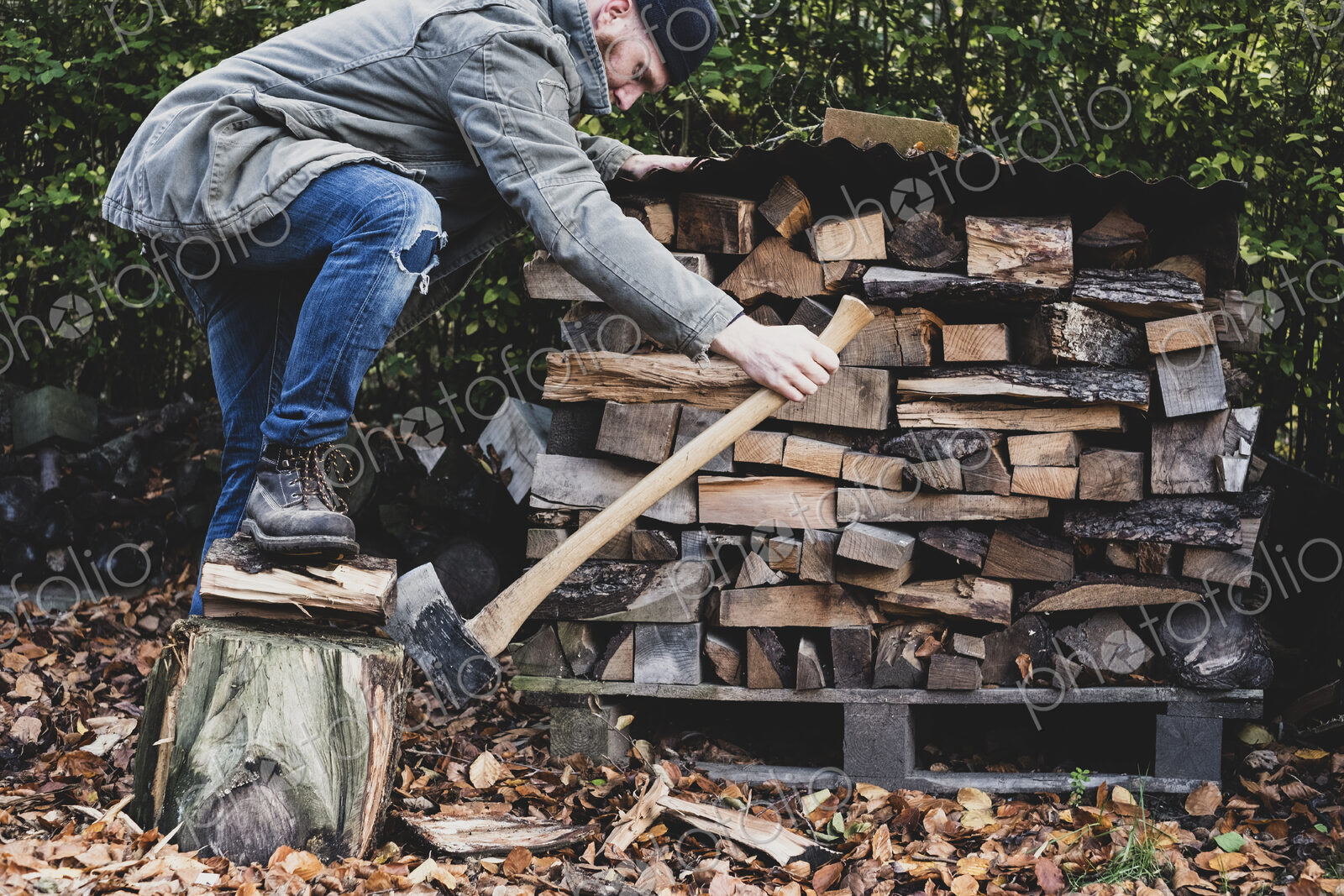 Bearded man wearing black beanie and parka standing in garden in autumn, using axe to…