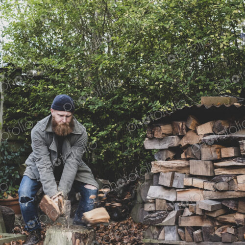 Bearded man wearing black beanie and parka standing in garden in autumn, using axe to…