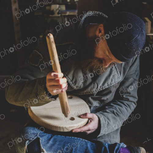 Bearded man wearing black beanie sitting in workshop, fitting leg to wooden stool.