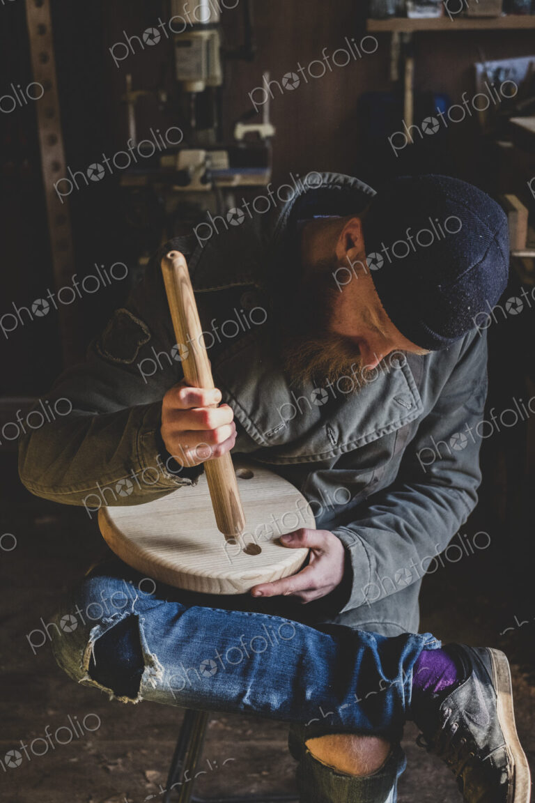 Bearded man wearing black beanie sitting in workshop, fitting leg to wooden stool.