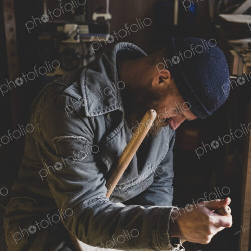 Bearded man wearing black beanie sitting in workshop, fitting leg to wooden stool.