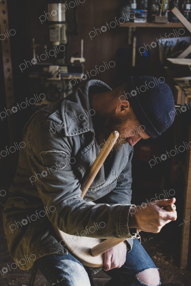 Bearded man wearing black beanie sitting in workshop, fitting leg to wooden stool.